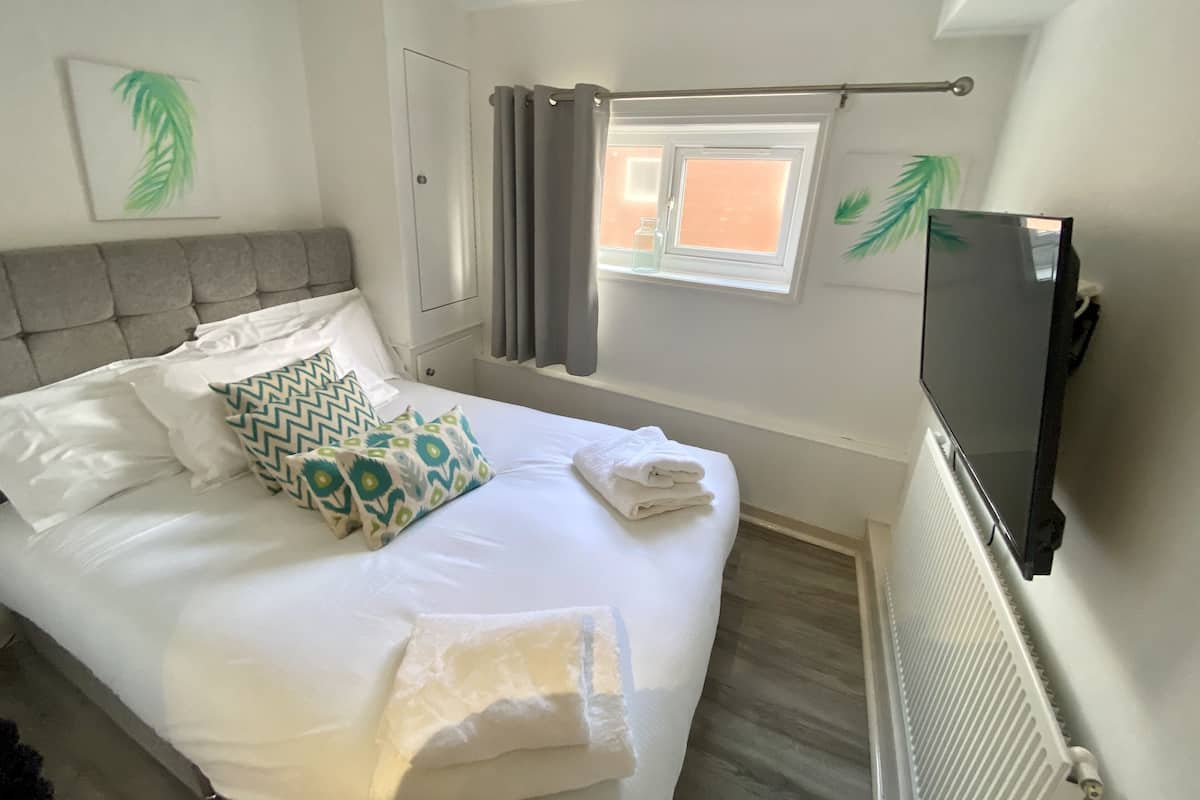 A neatly made bed with pillows, a window with curtains, a radiator, and a flat-screen TV mounted on the wall.