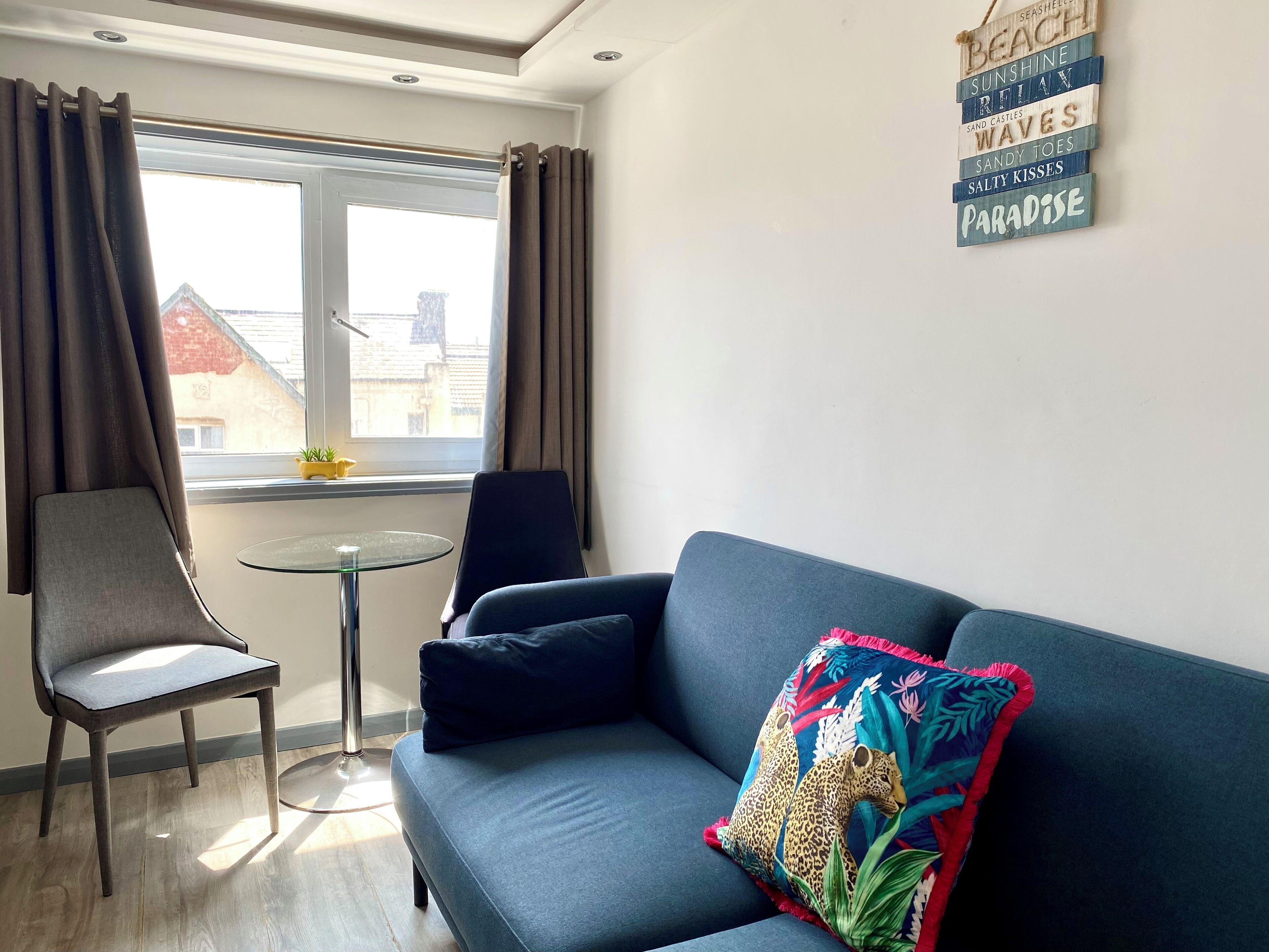 A modern living room with a blue sofa, a grey armchair, and a small round table. A window with curtains is visible, and a wall decoration with beach-related words is present.