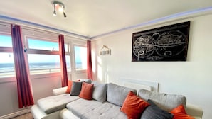 Ein modernes Wohnzimmer mit einem grauen Ecksofa, roten und grauen Kissen, einem großen Fenster mit Meerblick und einer Weltkarte an der Wand.