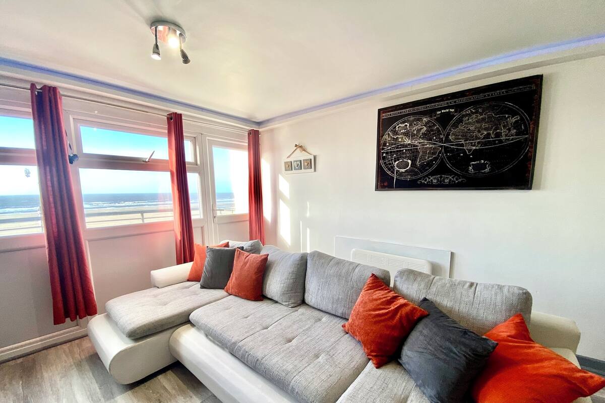 A modern living room with a grey sectional sofa, red and grey cushions, a large window with a view of the ocean, and a world map on the wall.