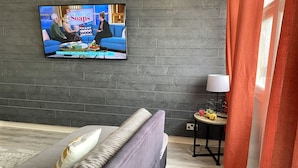 Ein modernes Hotelzimmer mit grauer Ziegelwand, einem Fernseher über einer Couch und einem kleinen runden Tisch mit Lampe und Dekoration.