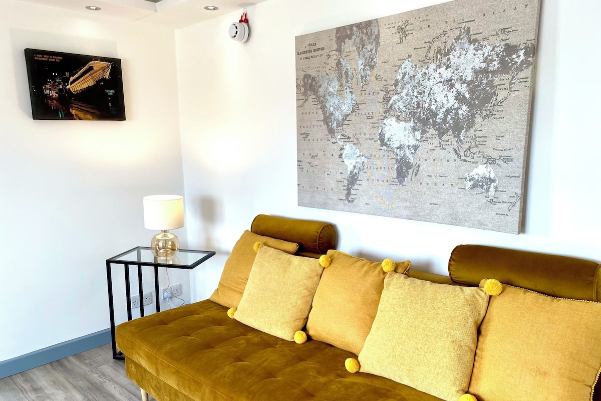 A room with a mustard-colored sofa, a small side table with a lamp, and a world map on the wall.