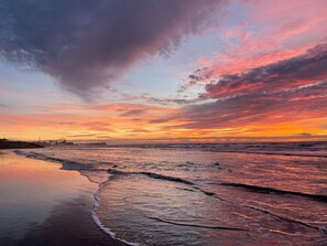 Une plage avec des vagues au coucher du soleil, une jetée au loin et un ciel nuageux.