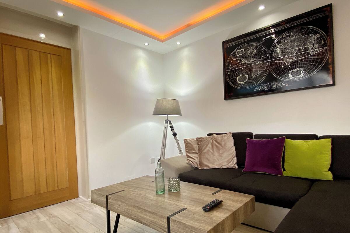 A modern living room with a wooden coffee table, a lamp, a black sofa with colorful cushions, and a world map artwork on the wall.
