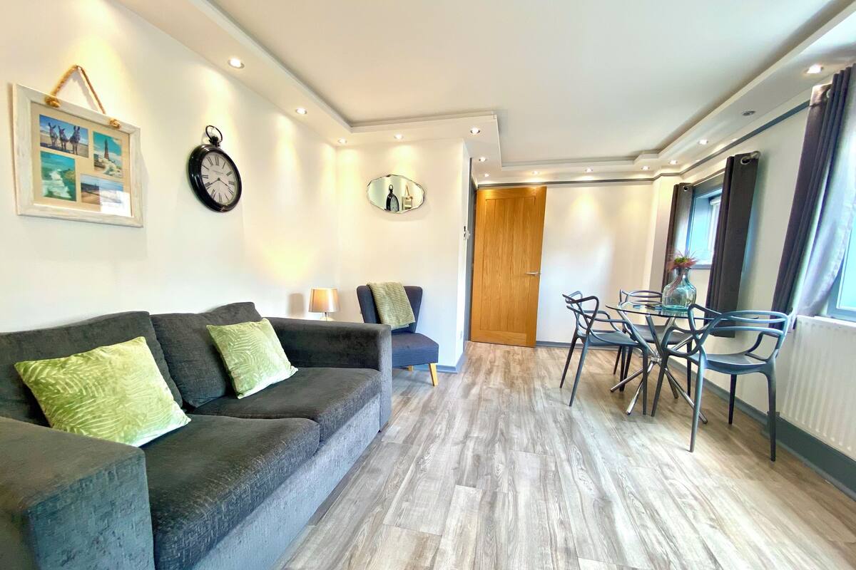A modern living room with a grey sofa, a dining table with chairs, a wooden door, and a clock on the wall.
