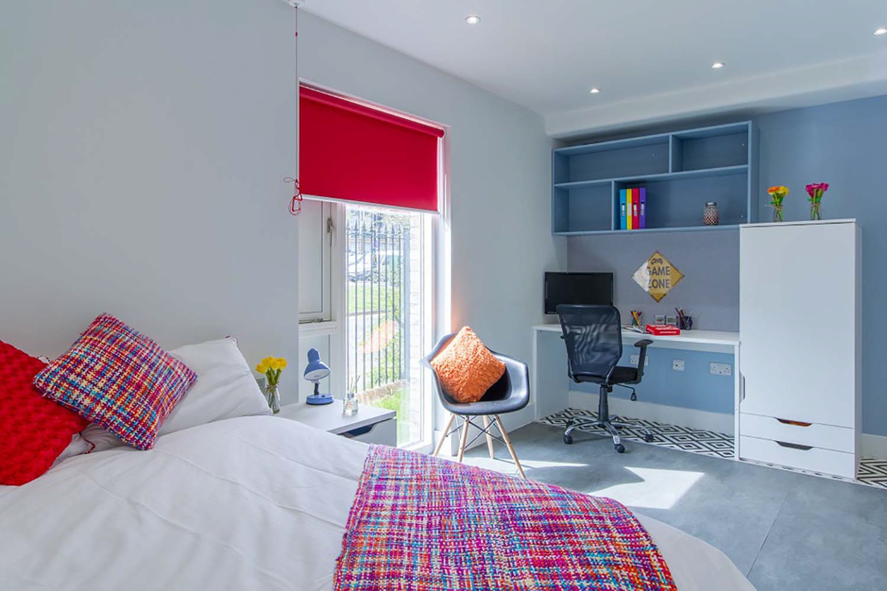 A bedroom with a bed, a desk, and a chair. There is a window with a red blind, a bookshelf, and a small table with a lamp.
