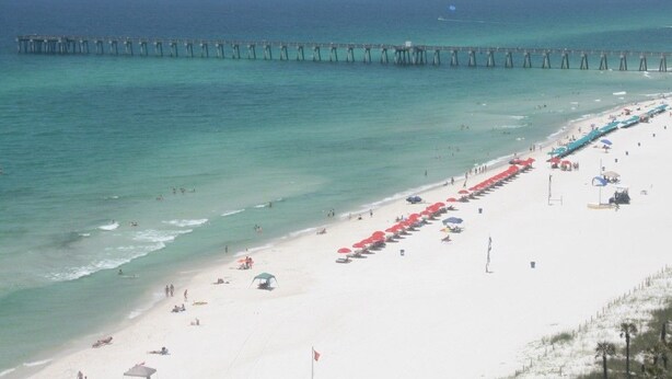 A beach with a pier extending into the ocean, rows of umbrellas, and people enjoying the seaside. - Clam Shell at Grand Panama - Intimate and Serene Perfect spot for relaxing (Panama City Beach)