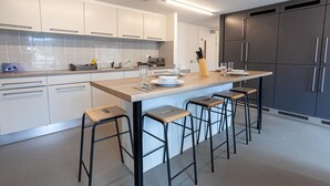 A modern kitchen with white cabinets, a central island with stools, and a dark refrigerator. - Axo Paradise village (Coventry)