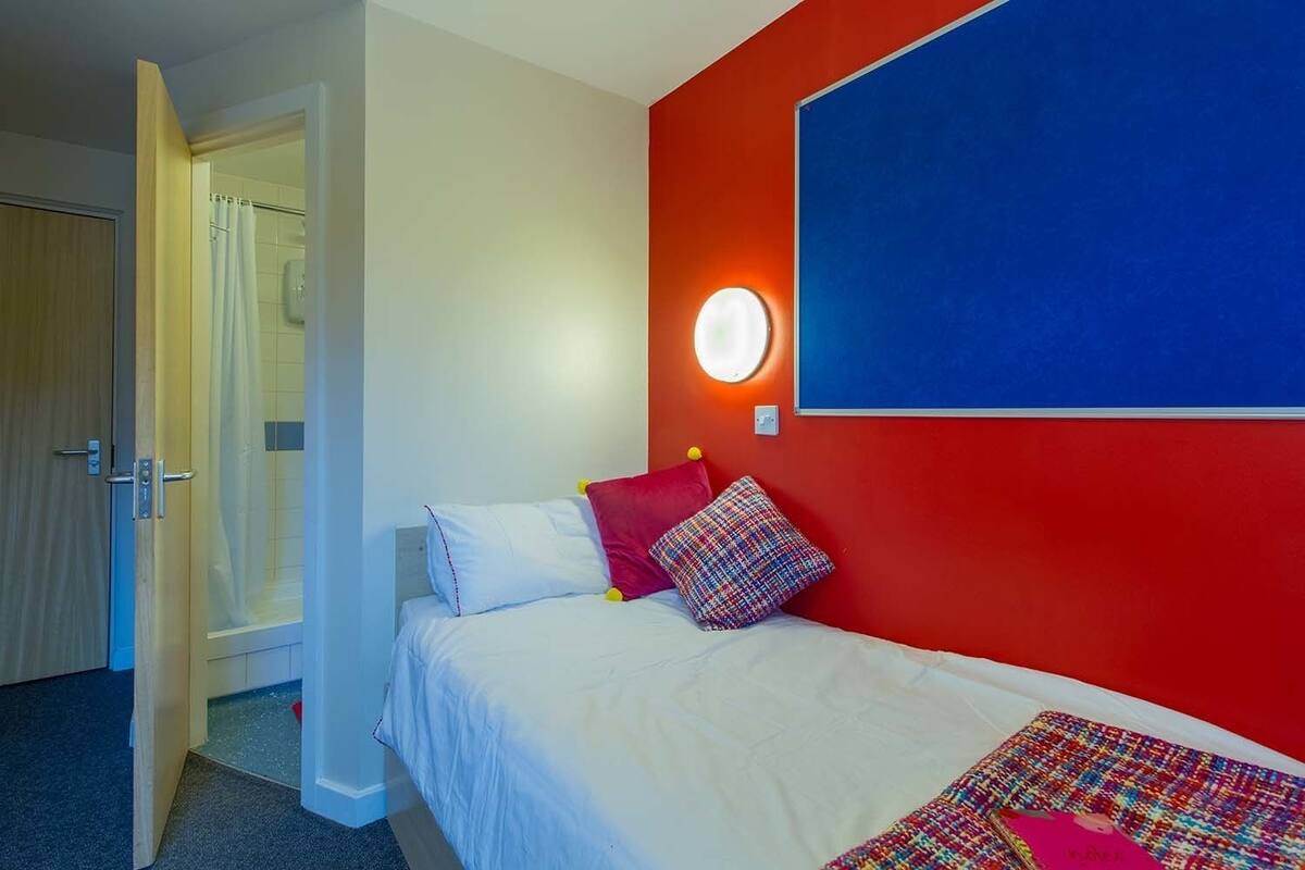 A hotel room with a bed, a red accent wall, a round wall light, and a door leading to a bathroom.