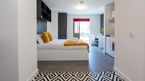 A modern hotel room with a bed, a desk, a kitchenette, and a window with a red frame. - Axo Paradise village (Coventry)