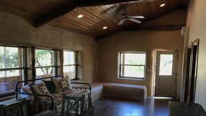 A rustic room with a wooden ceiling, a bed with patterned bedding, a bench, and a window with a view of trees. - Cabin 4 of 7 With Private River Access to the Nueces River (Uvalde)