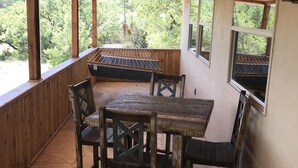 A covered outdoor dining area with a wooden table and chairs, large windows, and a view of greenery. - Cabin 4 of 7 With Private River Access to the Nueces River (Uvalde)