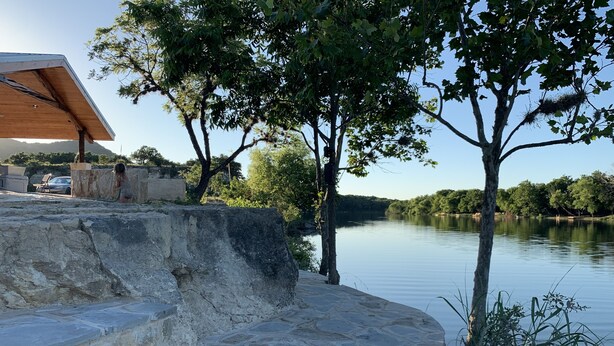 A stone staircase leading to a wooden deck overlooking a calm lake, surrounded by trees and a clear sky. - Cabin 4 of 7 With Private River Access to the Nueces River (Uvalde)