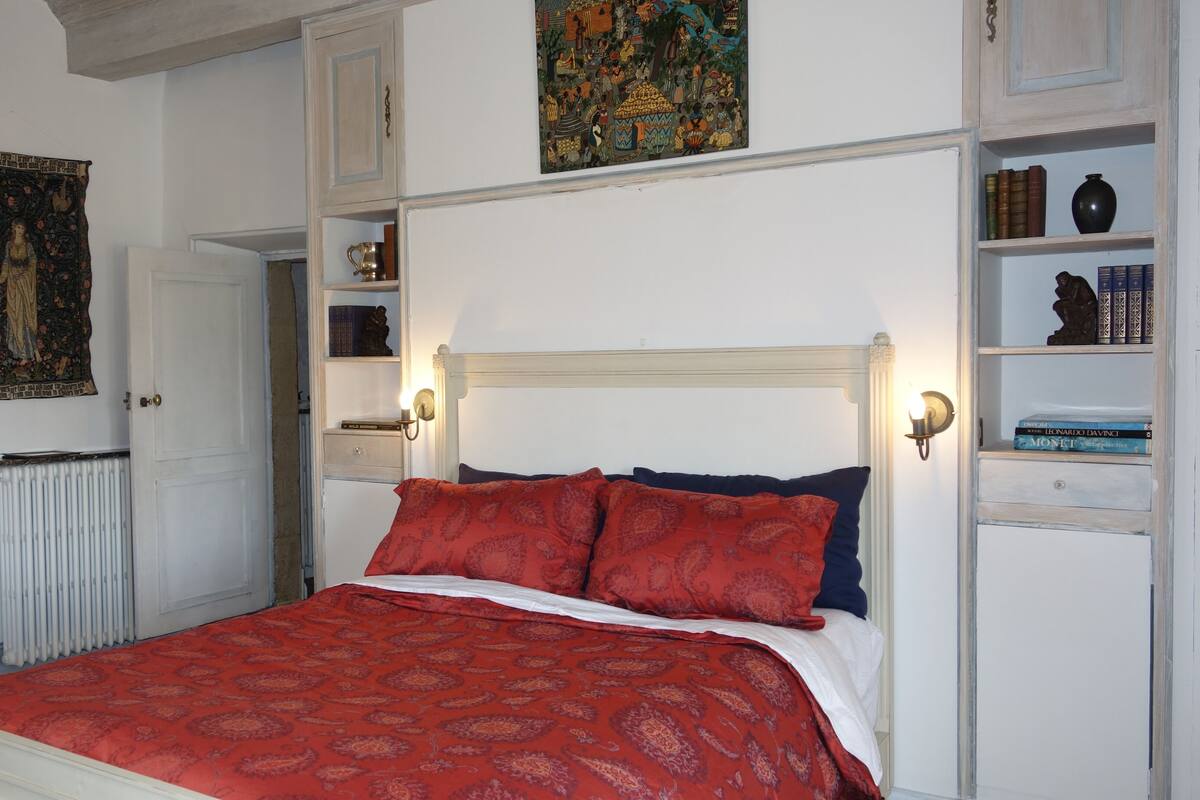A bedroom with a bed, a radiator, a wardrobe, a shelf with books, and a tapestry.