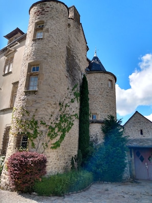 A historic stone tower with a turret and a window, surrounded by greenery and a paved area. - Château de Balsac (Druelle Balsac)