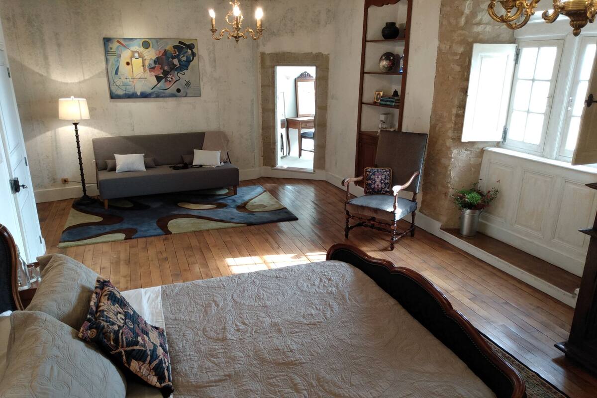 A spacious room with a large bed, a sofa, a chair, and a wooden floor. There is a painting on the wall and a chandelier.