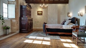 Quarto com piso de madeira, cama com peseira escura, cômoda de madeira e lustre.