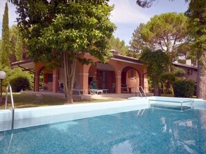 A swimming pool with a clear blue water surface, surrounded by a brick building with arched windows and a covered patio area. - Villa Sabrina (Lignano Sabbiadoro)