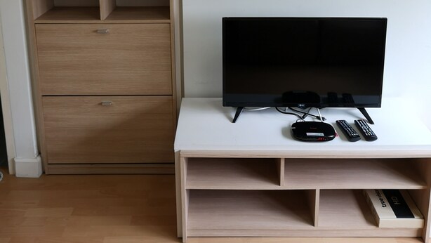 A room with a wooden wardrobe, a white TV stand with a flat-screen TV, remote controls, and a power strip. - Comfy Deluxe One Bedroom (Bangkok)
