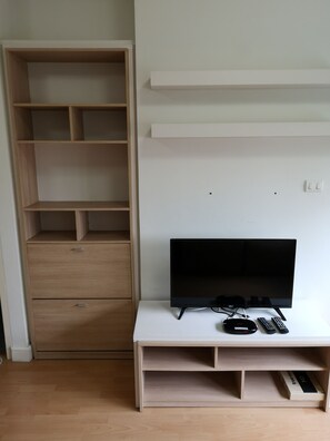 Una habitación con un armario de madera, un mueble blanco para televisión con un televisor de pantalla plana, controles remotos y una regleta eléctrica.