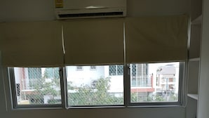 A room with a wooden floor, a window with a view of buildings, an air conditioning unit, and a white bench. - Comfy Deluxe One Bedroom (Bangkok)