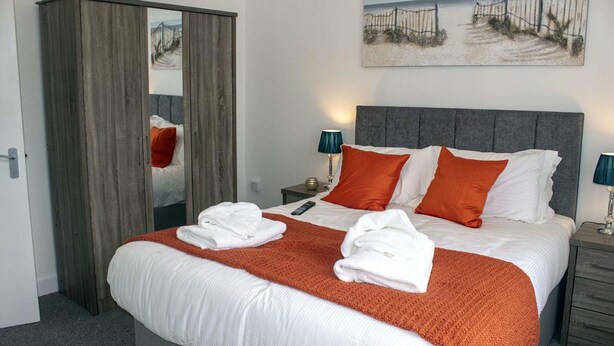 Una cama bien hecha con ropa de cama blanca y naranja, un cabecero gris y un cuadro encima.