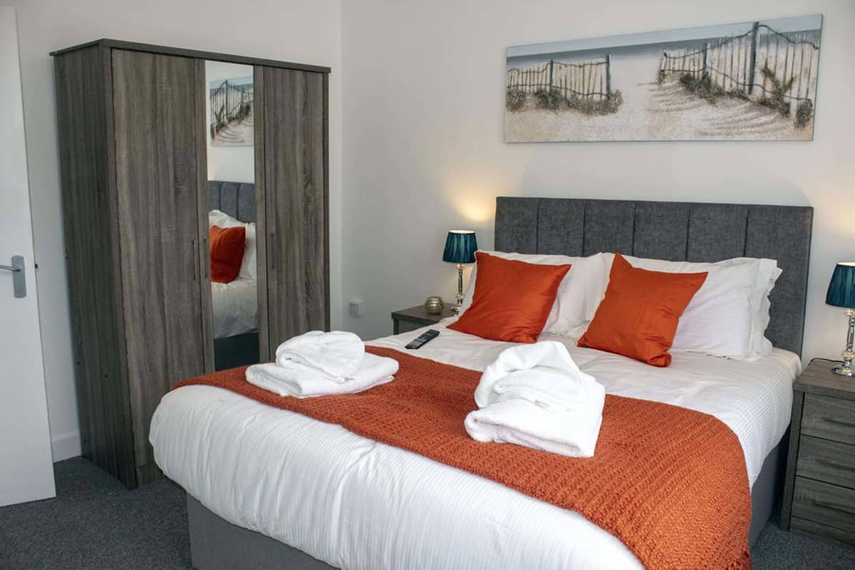 A neatly made bed with white and orange bedding, a grey headboard, and a painting above it.