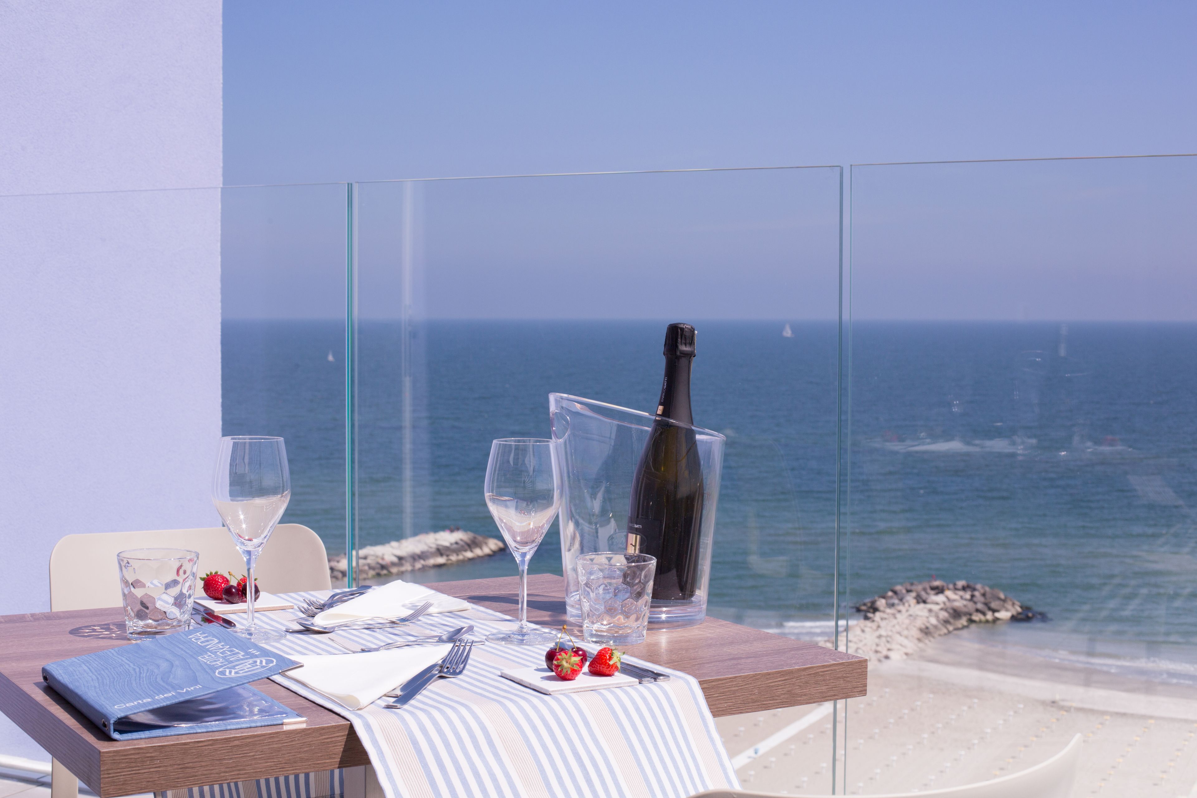 Una mesa dispuesta con una botella de champán, copas de vino y un menú en un balcón con vistas al mar.