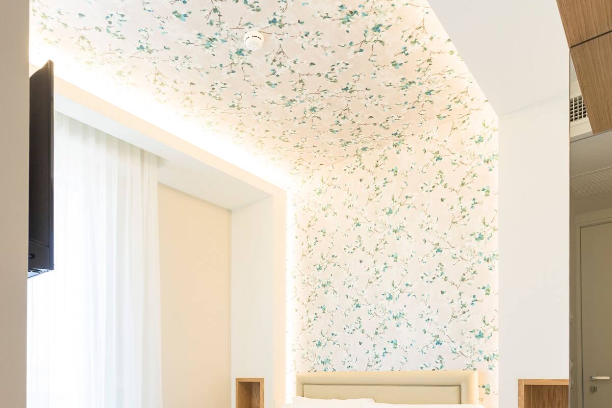 A bedroom with a bed, a wooden nightstand, a wardrobe, and a ceiling with floral wallpaper.