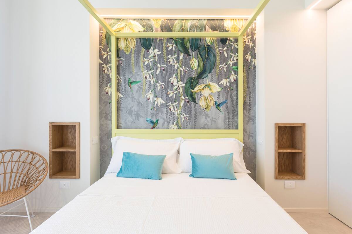 A bedroom with a bed, two pillows, a wooden chair, and a wall with a floral mural.