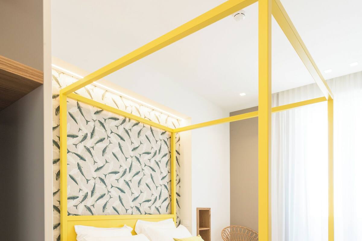 A bed with a yellow frame and white bedding, a patterned wallpaper, and a wooden chair in a modern room.