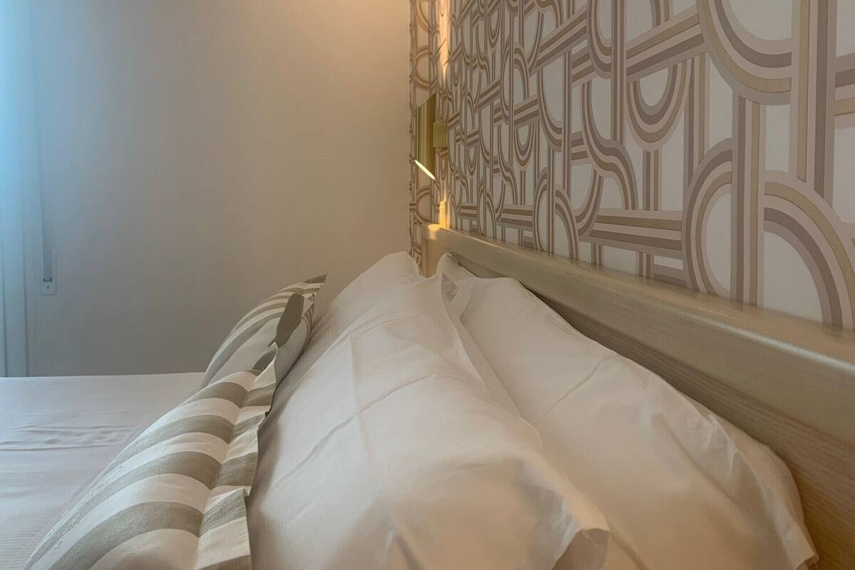 A neatly made bed with white and striped bedding, a decorative headboard with a geometric pattern, and a wall-mounted light fixture.
