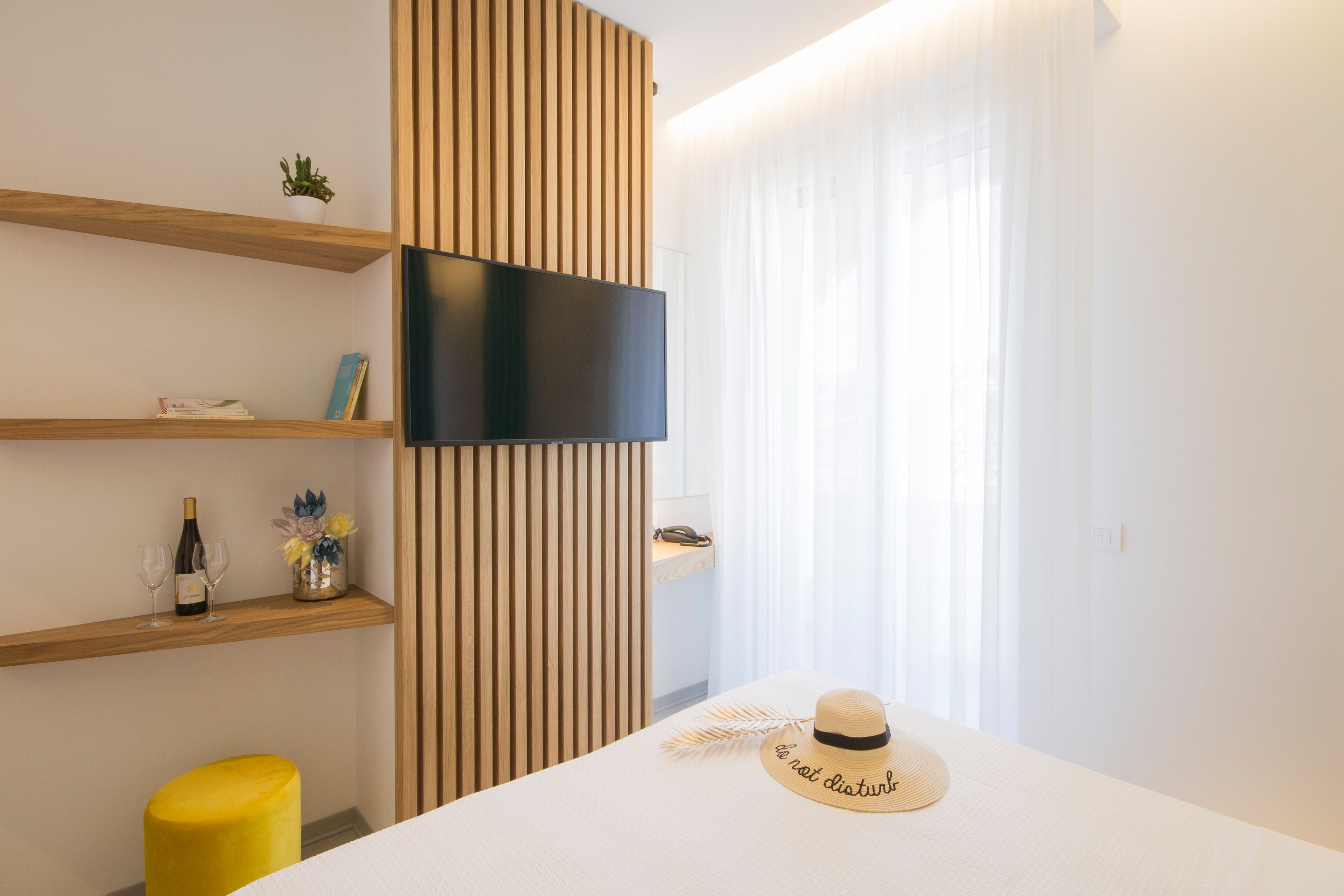 Hotelkamer met een flatscreen televisie aan de muur, houten planken met boeken en een fles, een geel poefje, en een bed met een strohoed.