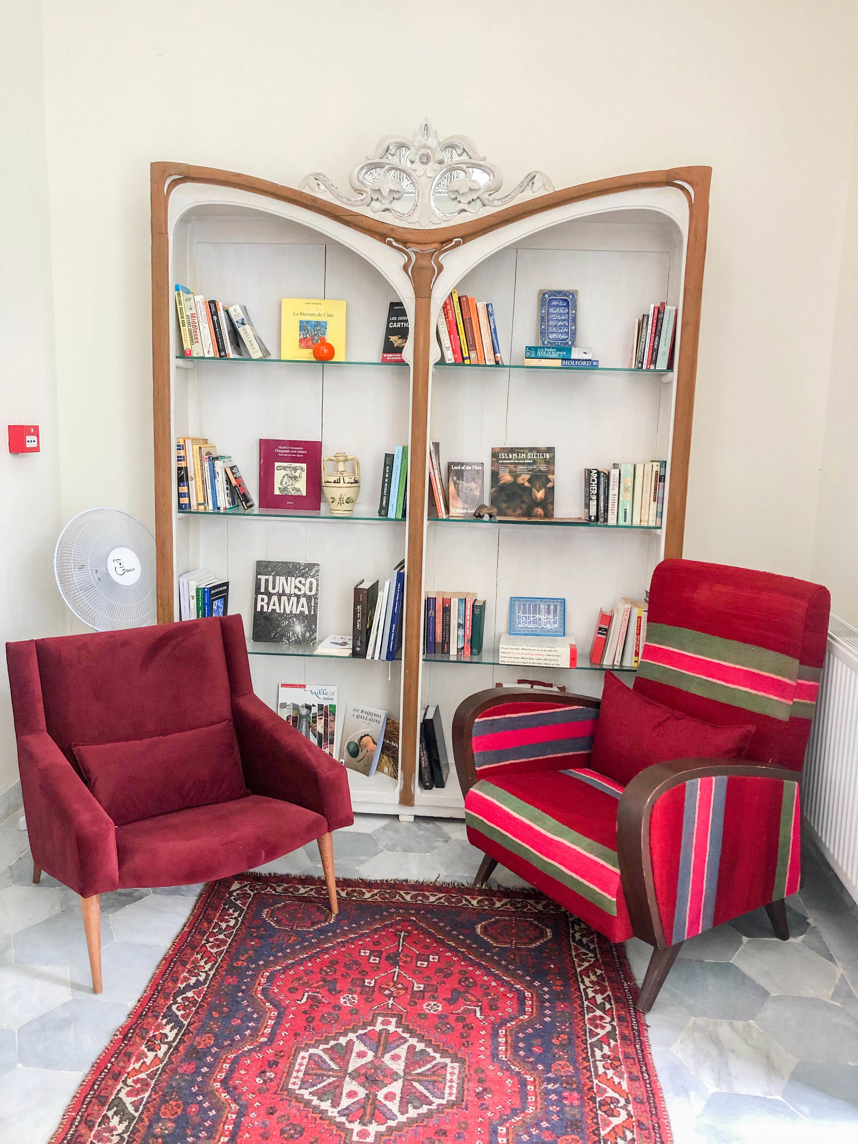 Una habitación con una amplia estantería abierta llena de libros, dos sillones, un tapete estampado y un radiador.