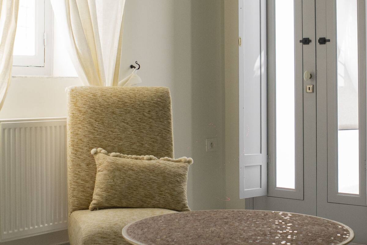 A room with a beige armchair, a round table, a radiator, a window with sheer curtains, and a door with a handle.