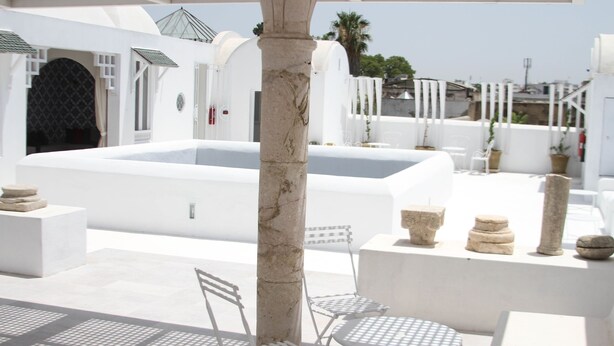 Une terrasse extérieure blanche, avec une piscine, des colonnes en pierre et des meubles en métal.