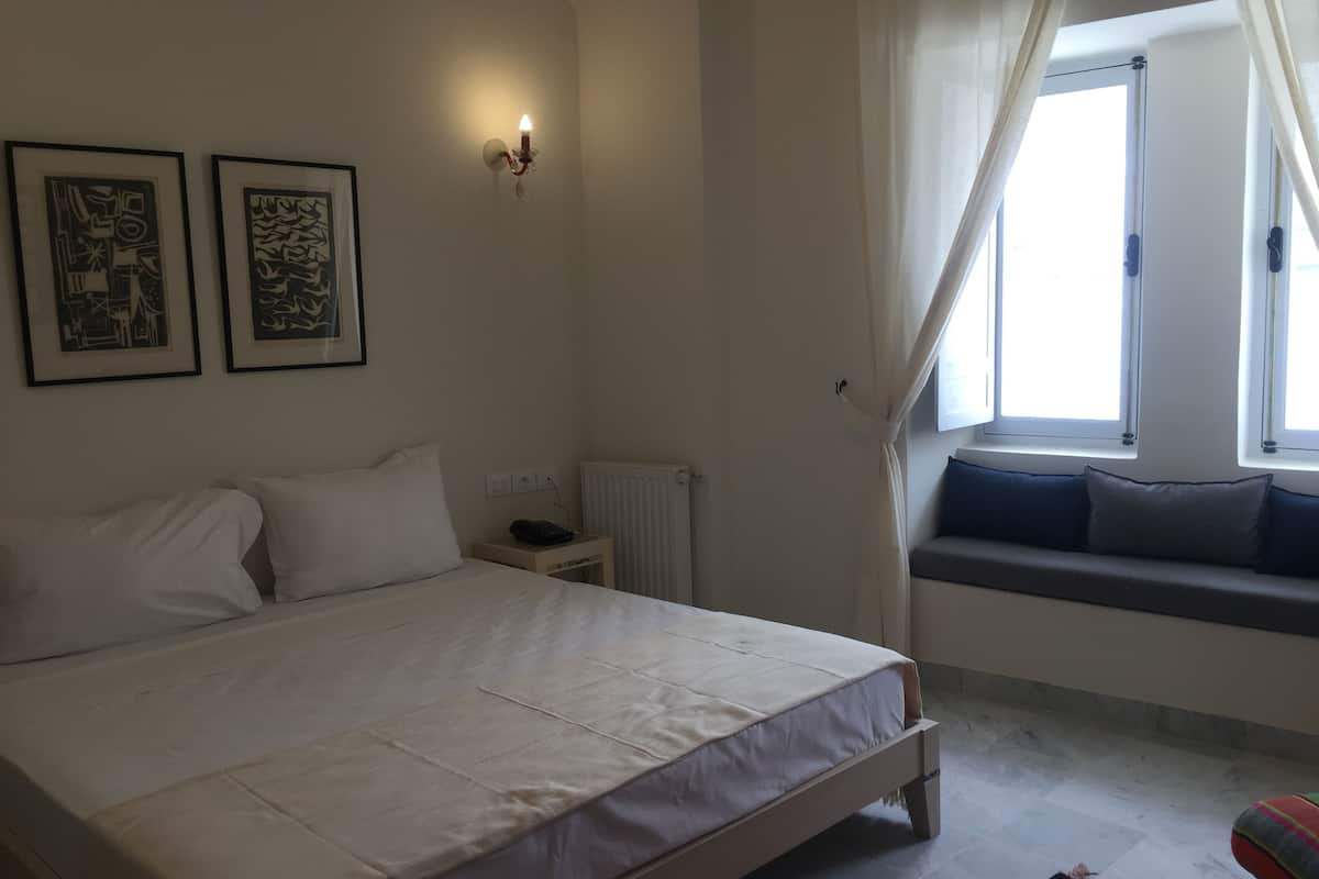A bedroom with a bed, a bedside table, a radiator, a window with curtains, and two framed pictures on the wall.