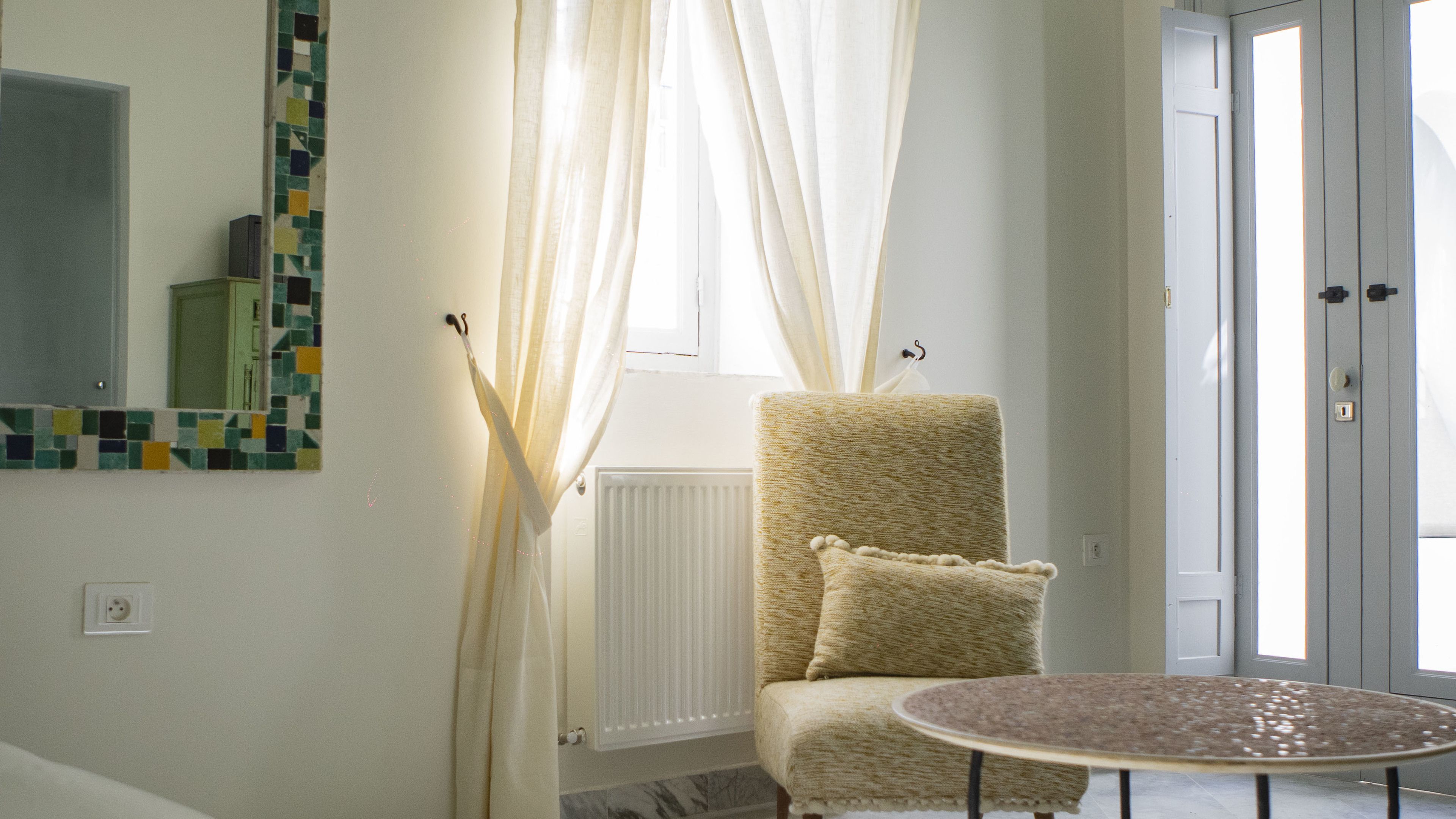 A room with a chair, a table, a window with curtains, a radiator, and a tiled wall.
