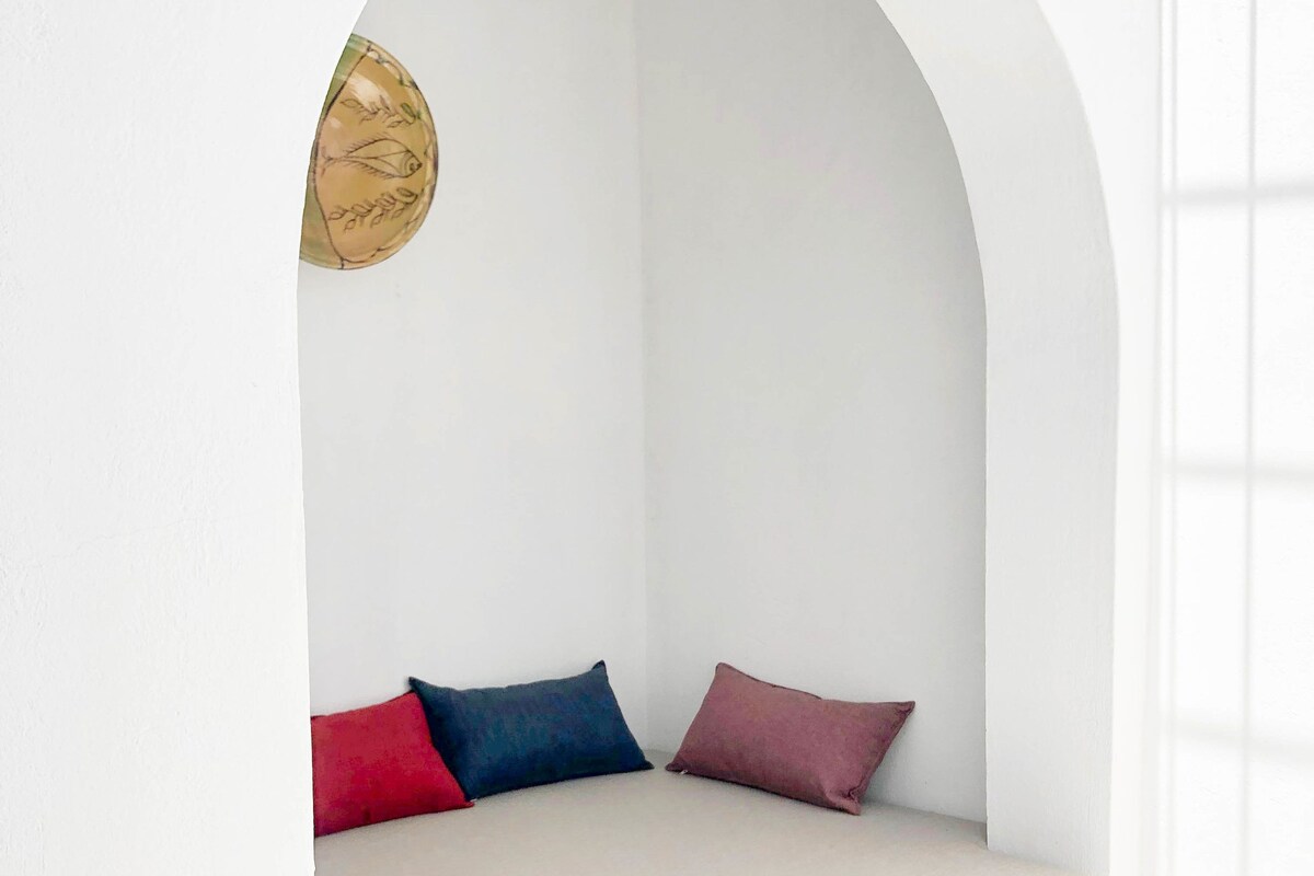 Una habitación minimalista con pared blanca, una ventana pequeña de arco y un banco acolchado con tres almohadas.