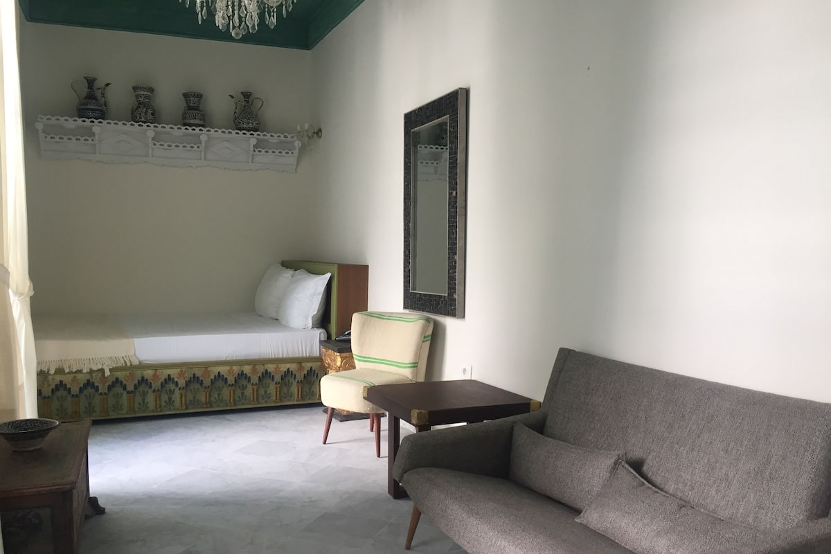 A room with a bed, a sofa, a chair, a small table, and a shelf with decorative items.