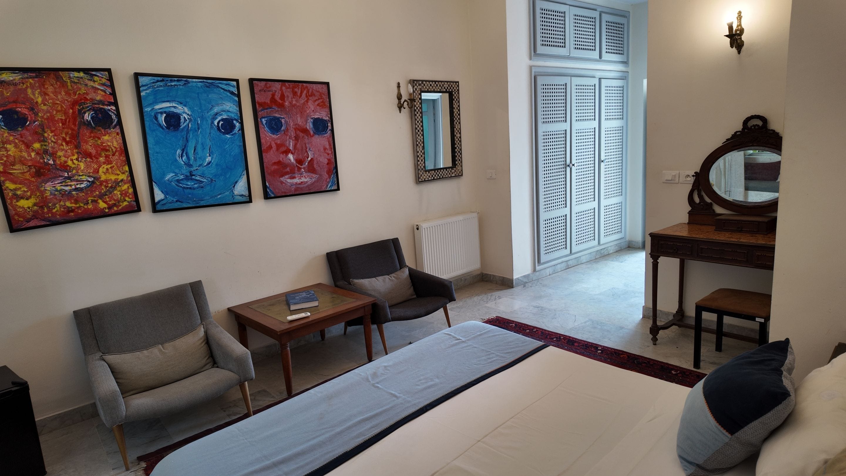 A hotel room with a bed, two armchairs, a small table, a dresser, a mirror, and three paintings on the wall.