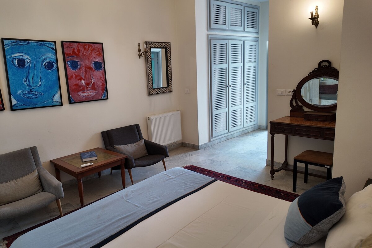 A hotel room with a bed, two armchairs, a small table, a dresser, a mirror, and three paintings on the wall.