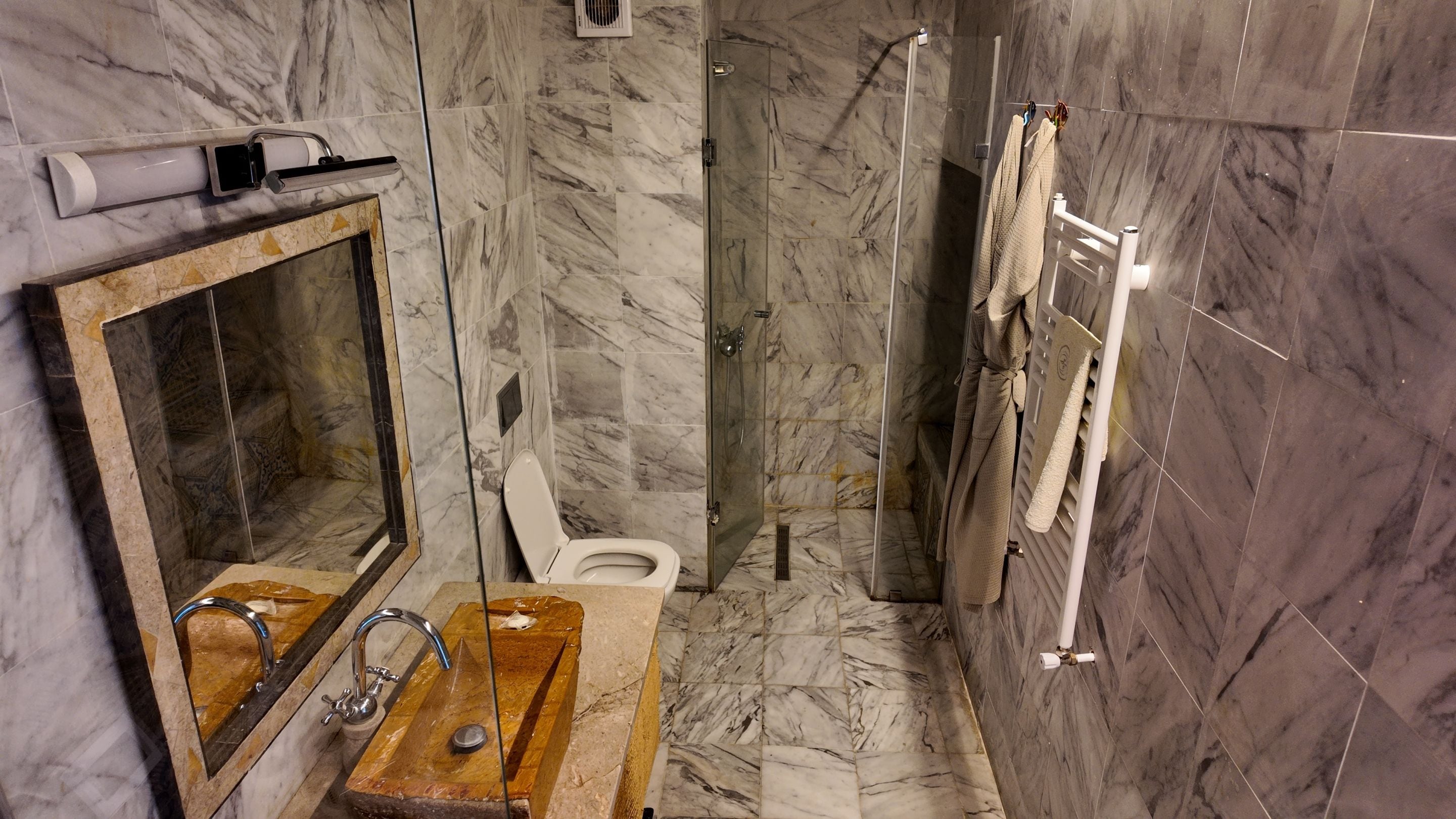 Een badkamer met een marmeren vloer, een houten wastafelmeubel met een wastafel, een toilet en een douche met glazen wanden.