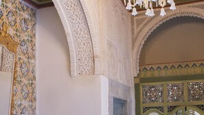 Une pièce richement décorée, avec un plafond haut, des motifs complexes sur les murs, un lustre, un banc, une table et un tapis à motifs.