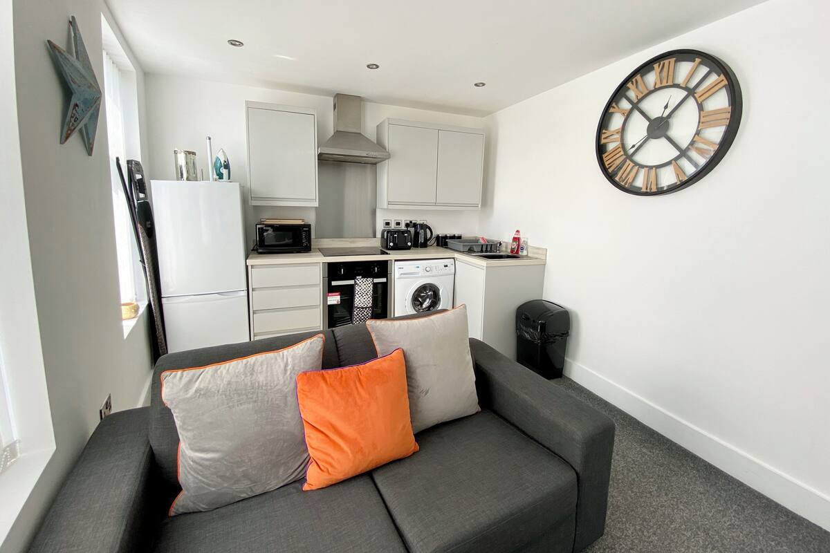 A compact living space with a kitchenette, a grey sofa with orange and grey cushions, and a large wall clock.