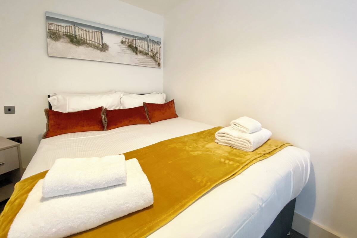 A neatly made bed with white bedding and a mustard-colored throw blanket, a painting on the wall, and a folded towel on the bed.