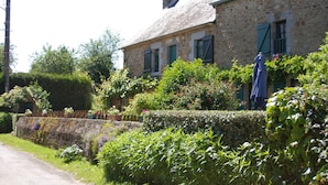 A stone building with a sloped roof and green shutters, surrounded by lush greenery and a stone wall. - Les Keriaden's Maison d'hotes (Pleudihen-sur-Rance)
