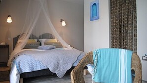 A bedroom with a bed under a mosquito net, a chair with a blue and white striped blanket, and a wall with a blue painting and a patterned curtain. - Les Keriaden's Maison d'hotes (Pleudihen-sur-Rance)