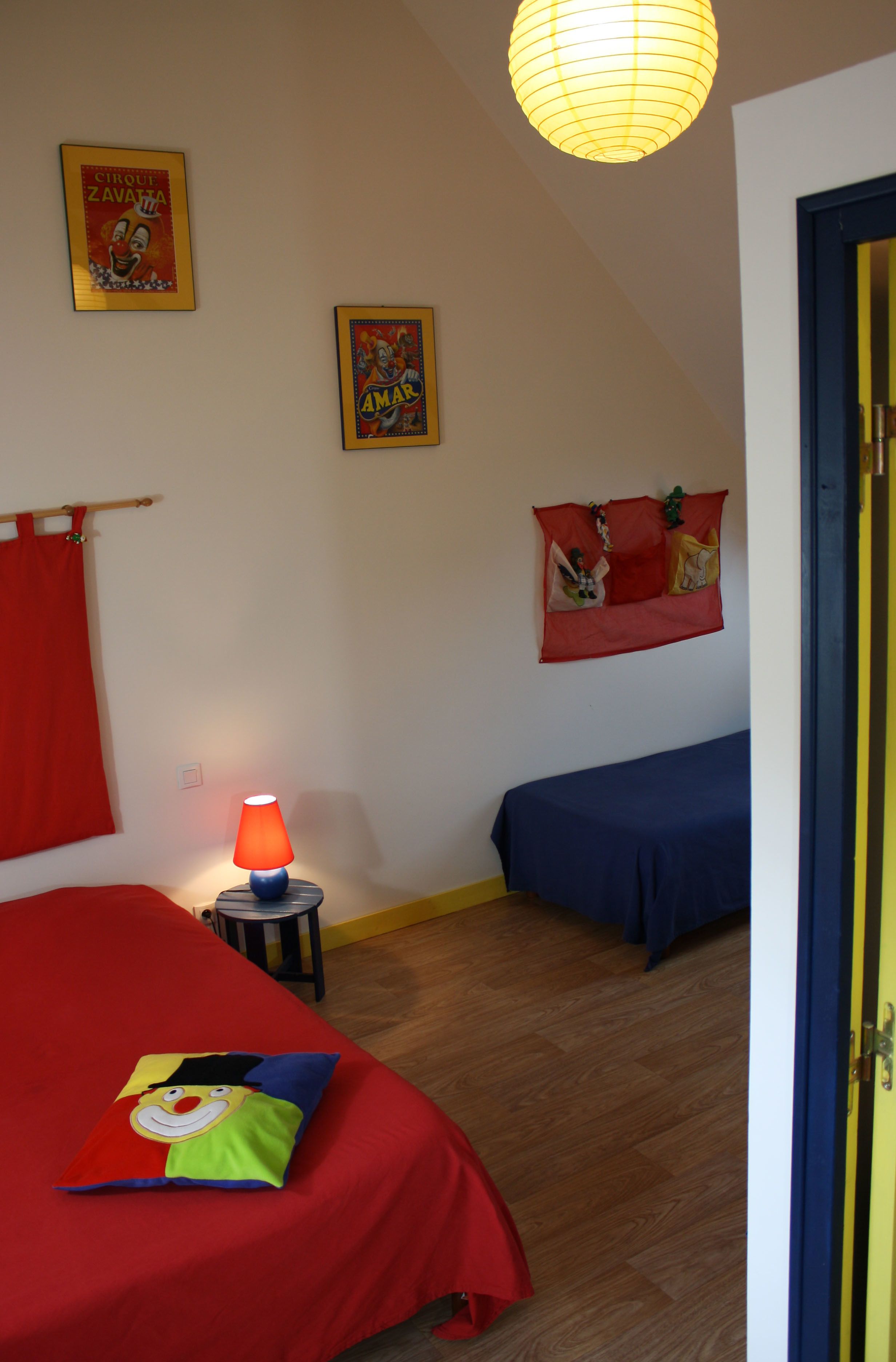 A room with two beds, one with a red bedspread and the other with a blue bedspread. There is a lamp on a small table next to the red bed, and two posters on the wall.