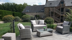 An outdoor patio with wicker furniture and a glass-top table, surrounded by a stone building and a wooden staircase. - Les Keriaden's Maison d'hotes (Pleudihen-sur-Rance)